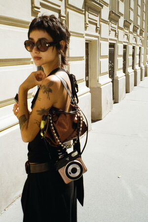 Eine Person mit kurzen dunklen Haaren, Sonnenbrille und sichtbaren Tattoos steht im Freien in der Nähe eines beigen Gebäudes und trägt einen INA KENT-Rucksack und eine Kamera.