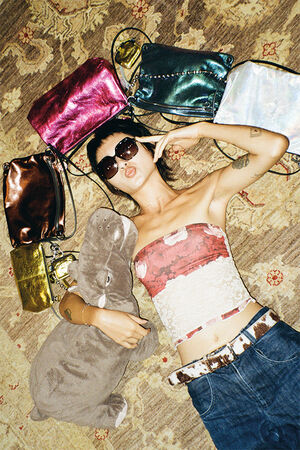 Eine Frau mit Sonnenbrille und trägerlosem Oberteil liegt auf einem gemusterten Teppich, hält ein ausgestopftes Nilpferd und ist von bunten INA KENT-Handtaschen umgeben.