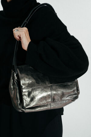 Eine schwarz gekleidete Person hält eine INA KENT Metallic-Leder-Umhängetasche mit einem schwarzen Riemen.