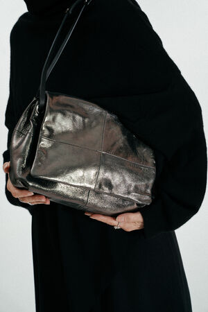 Eine schwarz gekleidete Person hält eine große, metallisch silberne INA KENT-Lederhandtasche unter dem Arm vor einem schlichten Hintergrund.