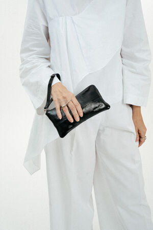 Eine Person, die ein lockeres weißes Outfit trägt, hält eine kleine schwarze INA KENT-Ledertasche in ihrer linken Hand vor einem unifarbenen Hintergrund.