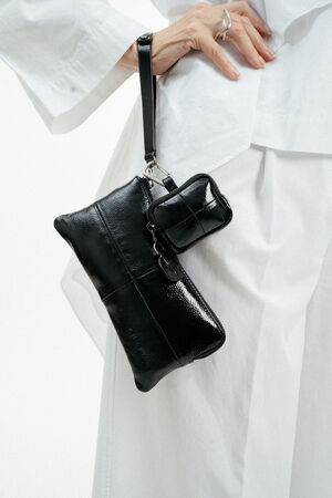 A person in a white outfit holds an INA KENT black patent leather wristlet bag with a small matching pouch attached.