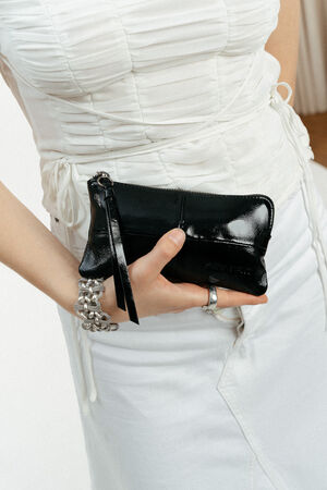 Person in a white ruched dress holds an INA KENT small black leather clutch with a zipper and wrist strap, featuring a chunky silver bracelet on their wrist.