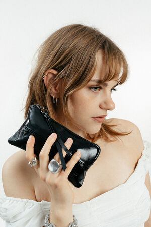 A woman with light brown hair in a white off-shoulder top holds an INA KENT small black zippered clutch near her face. She is accessorized with silver jewelry.
