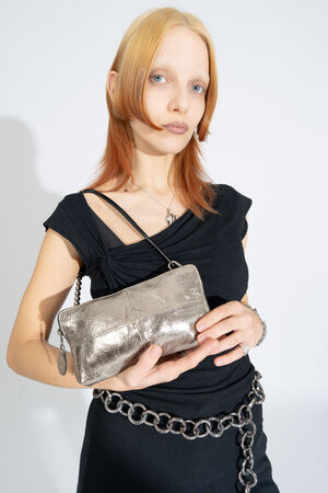 Person with light hair wearing a black outfit holds an INA KENT metallic silver clutch purse, with a chunky chain belt around their waist, standing against a plain white background.