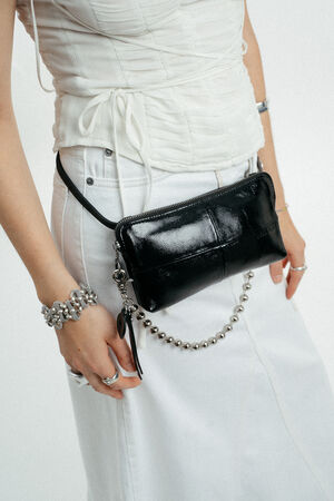 Eine Person, die ein weißes gerafftes Oberteil und einen Rock trägt und dazu eine schwarze INA KENT Crossbody-Tasche aus Lackleder, ein silbernes Armband und Ringe trägt.