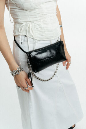 Eine Person trägt ein weißes Rüschenoberteil und einen Rock, dazu Silberschmuck und eine schwarze INA KENT Crossbody-Tasche aus Leder mit einem schicken Kettenriemen.