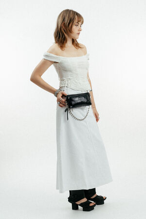 Eine Person, die ein schulterfreies weißes Oberteil, einen langen weißen Rock, schwarze Plateausandalen und eine schwarze INA KENT Crossbody-Tasche mit einem perlenbesetzten Riemen trägt, steht vor einem unifarbenen Hintergrund.