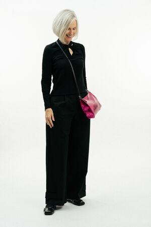 A woman with gray hair wearing a black top and wide-leg black pants stands against a white background, showcasing a bright pink INA KENT crossbody bag.