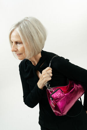 A woman with short gray hair wearing a black top holds a shiny pink INA KENT handbag over her shoulder against a plain white background.
