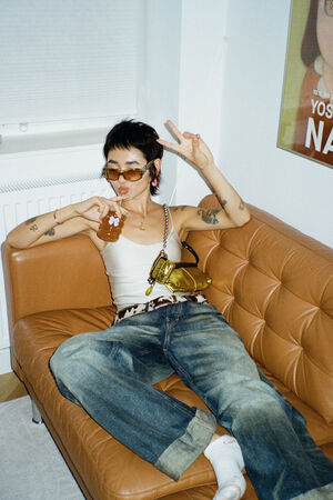 Person in sunglasses and casual clothes lounges on a tan sofa with an INA KENT bag by their side, holding a drink pouch and making a peace sign with one hand.
