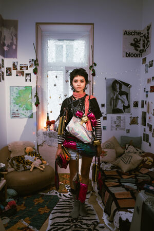 A young person stands in a cluttered, eclectic bedroom decorated with photos, posters, and string lights, wearing colorful, layered clothing and holding multiple shiny INA KENT bags.