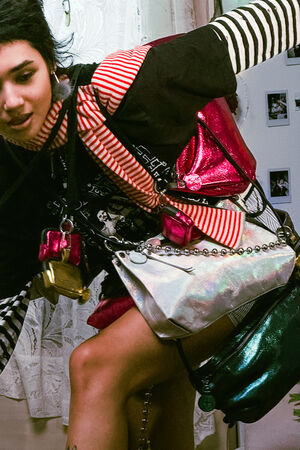 Person wearing multiple metallic INA KENT bags and a red-and-white striped scarf, with layered clothing, standing in a room with lace curtains and photos on the wall.