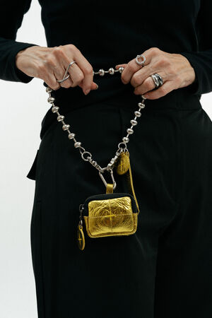 Person wearing a black outfit holds a beaded chain with an INA KENT metallic gold pouch and keychain attached, showcasing multiple silver rings on their fingers.