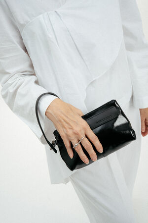 A person dressed in white holds a small black INA KENT leather clutch in their left hand, with a silver ring on their finger.