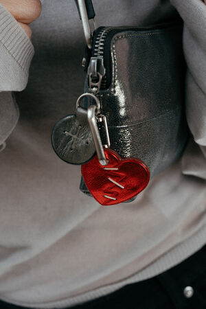 A close-up of a metallic INA KENT zippered pouch with a keychain featuring a red heart-shaped charm and a round gray tag attached.
