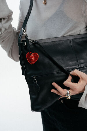 Person wearing a light long-sleeve top holds an INA KENT black leather bag with a red heart keychain attached. Only the torso and hands are visible.
