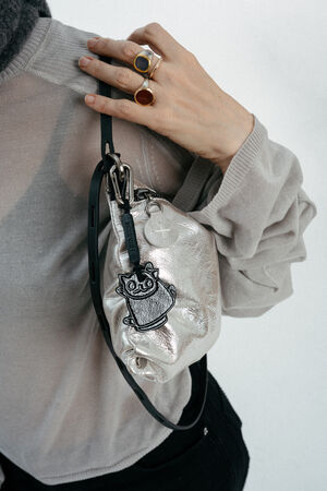 Person wearing multiple rings holds a small, metallic INA KENT bag with a black cat design and a round tag, held close to their shoulder.