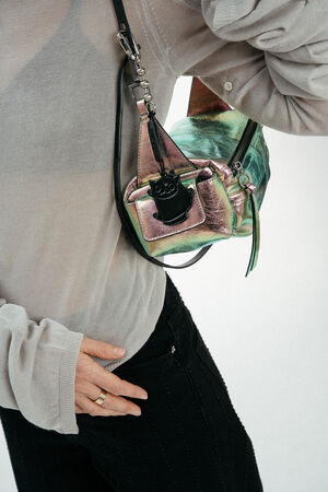 A person wearing a sheer, long-sleeve top and black pants carries an iridescent INA KENT crossbody bag with a small black accessory attached.