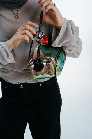A person wearing a gray top and black pants holds a shiny green INA KENT handbag adorned with a black and red heart-shaped keychain.