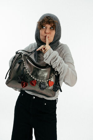 A person wearing a gray hood and sweater holds an INA KENT metallic handbag decorated with charms and gestures for silence with a finger to their lips.