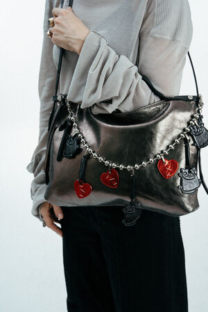 A person holds an INA KENT metallic handbag decorated with a chain of red heart and black cat charms. The person is wearing a light grey long-sleeve top and black pants.