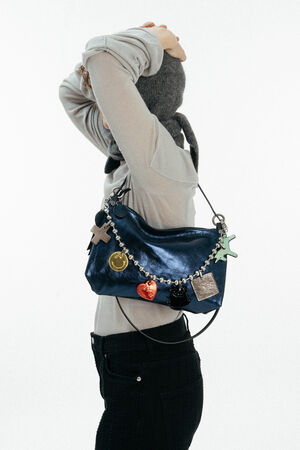 Person in a light long-sleeve shirt and gray headscarf poses with a shiny blue INA KENT handbag decorated with colorful charms over their shoulder.