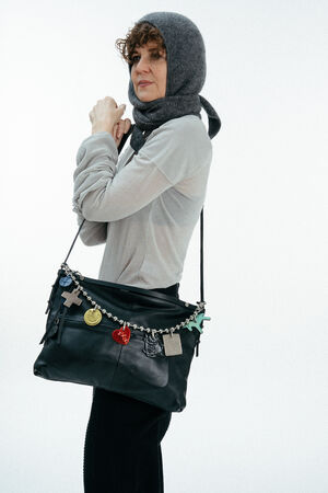 A person wearing a grey headscarf, light long-sleeve shirt, and dark pants carries an INA KENT black shoulder bag decorated with various colorful charms.