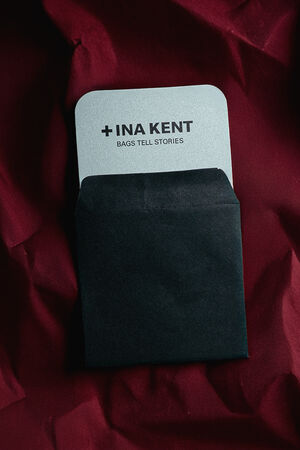A black envelope partially reveals an INA KENT card with the text "+INA KENT BAGS TELL STORIES," set against a dark red, textured background.