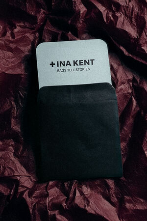 A card with the text "+INA KENT BAGS TELL STORIES" partially visible inside a black envelope, rests on crumpled dark fabric—a glimpse into INA KENT's world of storytelling accessories.