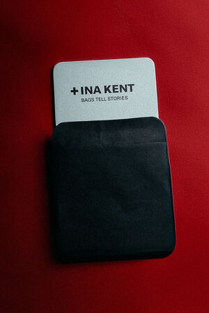 A silver INA KENT card with the text "+INA KENT BAGS TELL STORIES" partially slides out of a black envelope on a bold red background.