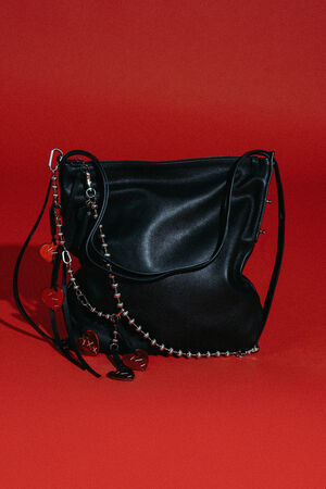 Eine INA KENT Handtasche aus schwarzem Leder mit Reißverschluss, Metallbeschlägen und dekorativen Anhängern vor einem einfarbig roten Hintergrund.