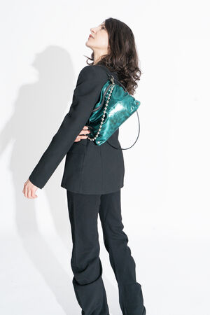 Eine Frau in einem schwarzen Anzug steht seitlich und blickt nach oben, wobei sie eine glänzende grüne INA KENT-Handtasche vor einem weißen Hintergrund über die Schulter wirft.