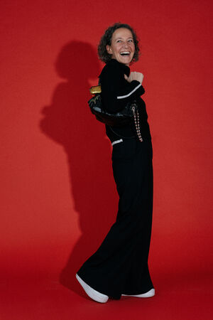 Eine Person mit kurzen lockigen Haaren, schwarzer Kleidung und weißen Schuhen steht lächelnd vor einem einfarbig roten Hintergrund und hält eine schwarze INA KENT-Tasche über der Schulter.