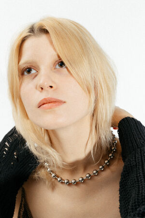 A person with shoulder-length blonde hair looks upward, wearing a chunky beaded necklace, a black sweater, and an INA KENT accessory against a plain white background.