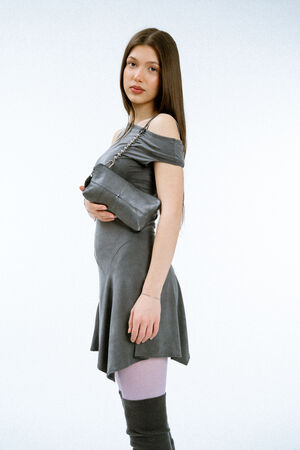 Weiblich gelesenes Modell in grauem Kleid mit einer kleinen Schultertasche aus Leder und schwarzem Riemen mit metallischen Kugelelementen