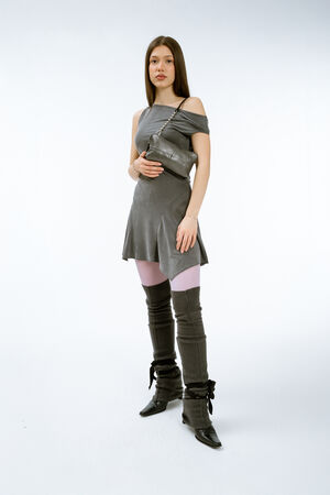 Weiblich gelesenes Modell in grauem Kleid mit einer kleinen Schultertasche aus Leder und schwarzem Riemen mit metallischen Kugelelementen