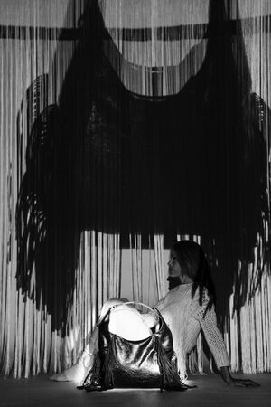 A woman sits on the floor beside an INA KENT handbag, with long fringe curtains and a large shadow of the bag cast on the wall behind her.
