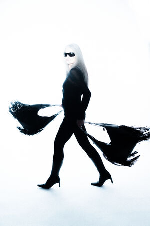 A person in black clothing, high-heeled boots, and sunglasses walks in a white space, holding two large black fringed INA KENT bags that trail behind them.