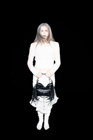 A woman in a white knit outfit stands against a black background, holding an INA KENT black fringe handbag with both hands, looking directly at the camera.