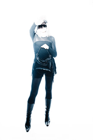 A person wearing sunglasses, a mask, dark clothing, and boots stands holding an INA KENT handbag with one arm raised, against a white background.