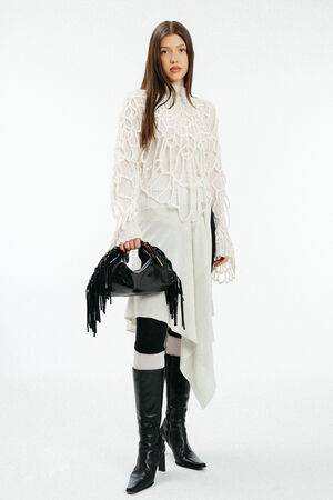 Weiblich gelesenes Model in losem, weißen Strick, mit schwarzer Ledertasche mit Fransen in der Hand