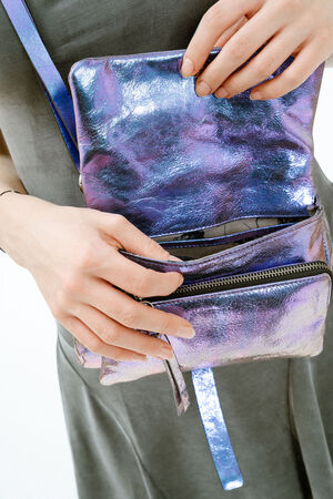 A person in a gray dress opens the zipper of a shiny, metallic purple INA KENT crossbody bag.
