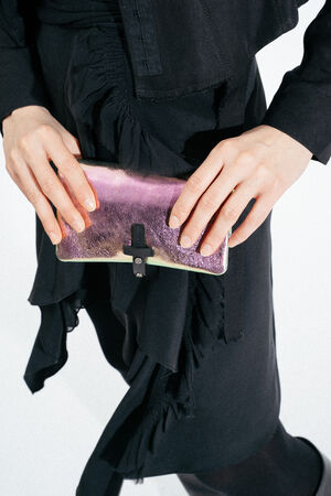 Eine schwarz gekleidete Person hält mit beiden Händen eine kleine, lila schillernde INA KENT-Clutch-Tasche.