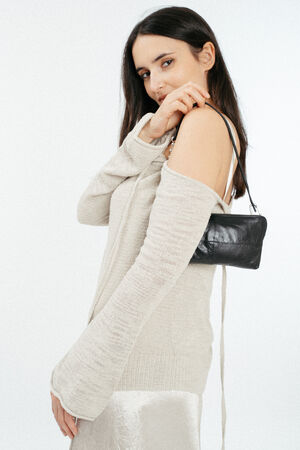 A woman wearing an off-shoulder beige sweater and a satin skirt holds a small black INA KENT shoulder bag against a plain white background.