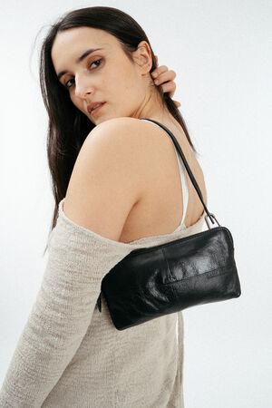 A woman with long dark hair poses in a beige off-shoulder sweater, holding an INA KENT black leather handbag on her shoulder against a plain white background.