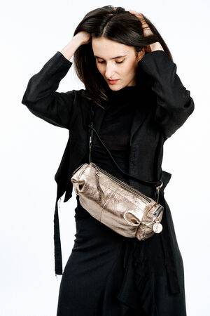 A woman in a black outfit stands against a white background, holding her hair, with an INA KENT metallic crossbody bag slung across her torso.