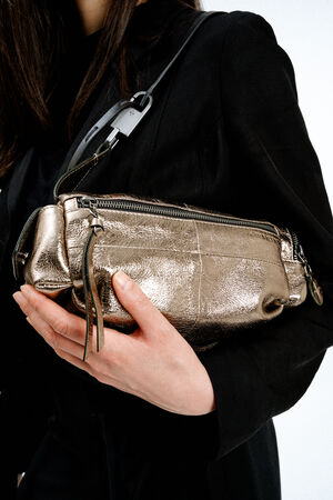 A person wearing a black outfit holds an INA KENT metallic gold leather handbag with zippers and a shoulder strap.