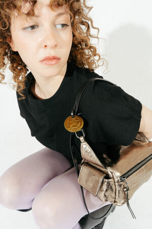 A woman with curly hair wearing a black top and light purple tights crouches, holding an INA KENT metallic gold handbag over her shoulder.