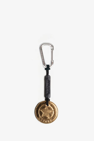 A keychain with a silver carabiner, black leather strap, and a round gold medallion featuring a star, the word "OSUJI," and crafted in the signature style of INA KENT.